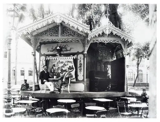Quiosque de chopp em parque, dono sentado, mesa vazia, placa com nome, estrutura aberta, árvores e prédio ao fundo.