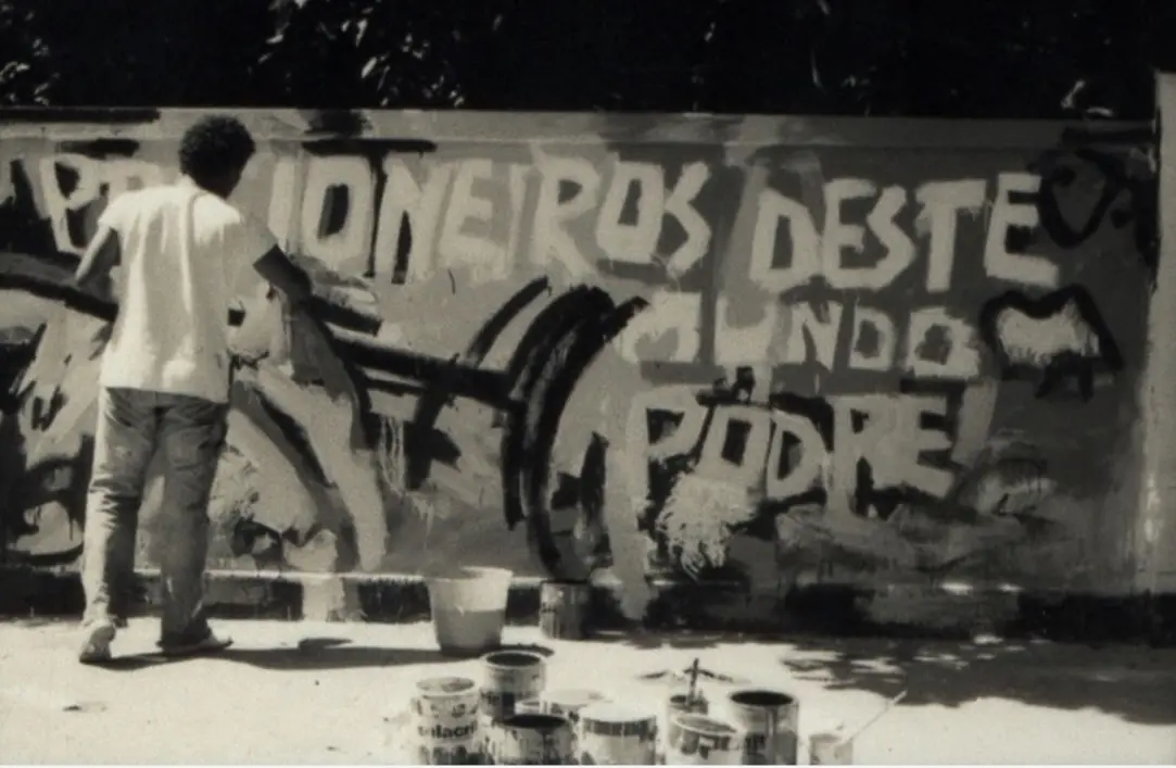 Homem pintando grafite em preto e branco, com palavras e elementos gráficos na parede.