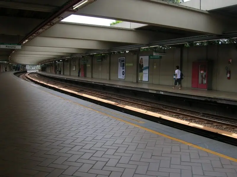 Plataforma de metrô com pessoa caminhando, azulejos cinza claro e placas informativas.