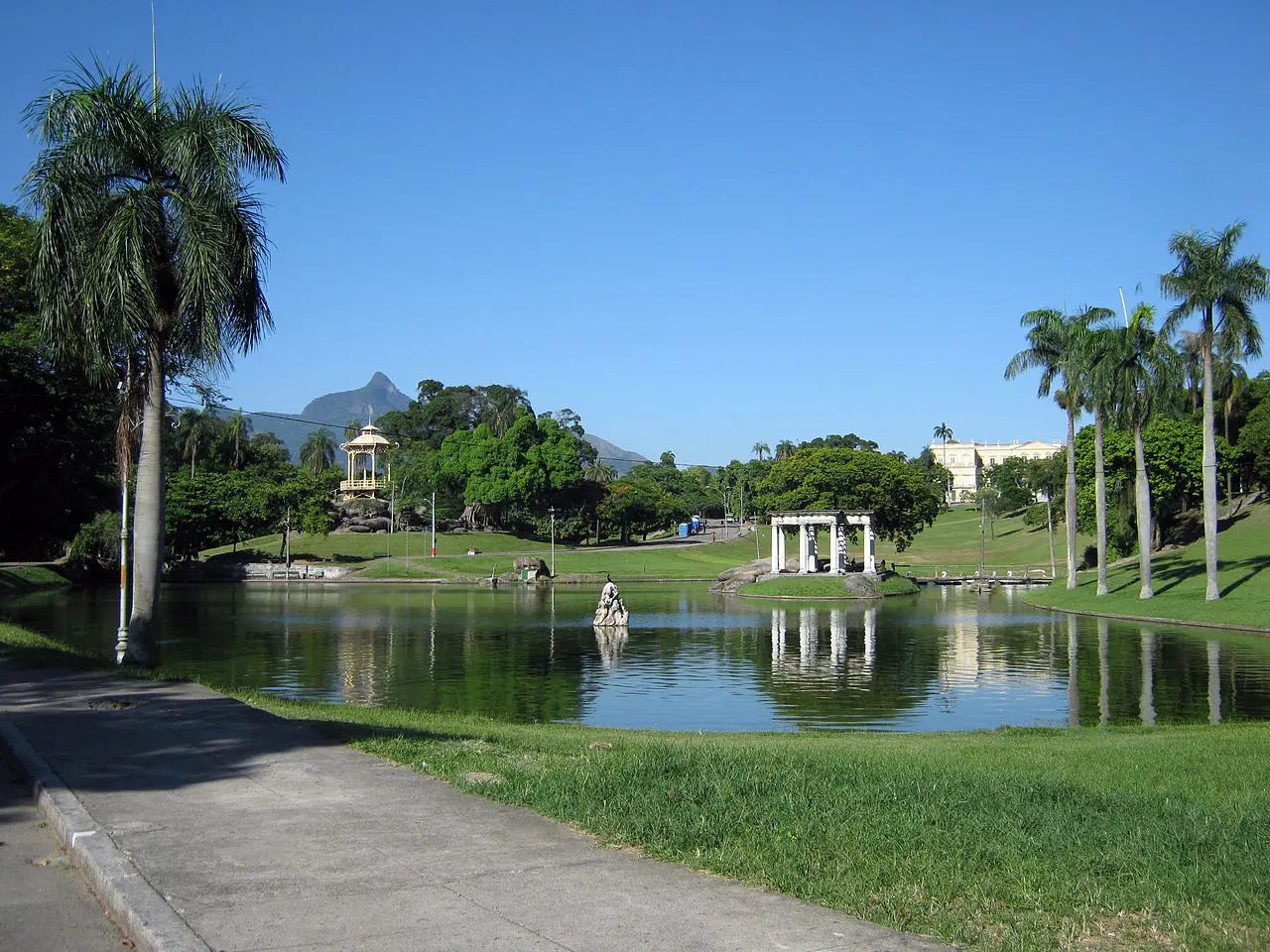 Parque com lago, palmeiras, estrada, kiosco, colina verde, estrutura arquitetônica, construção branca, árvore isolada, céu azul claro.