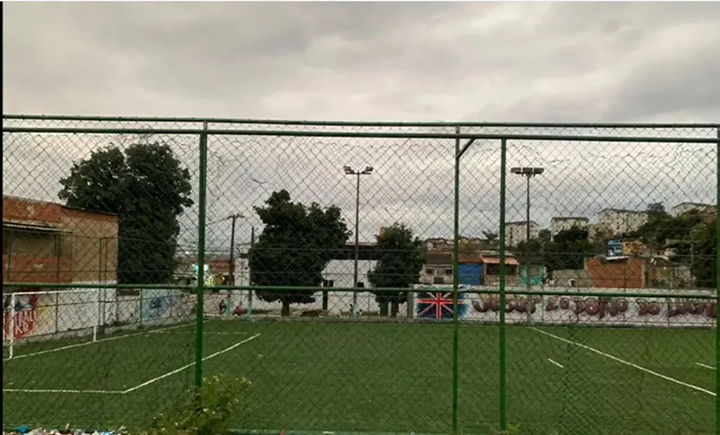 Campo de futebol cercado por grade verde, com construção marrom, árvores verdes e bandeira britânica.