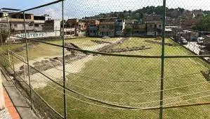 Campo de futebol cercado por rede, grama verde e construção ao fundo.