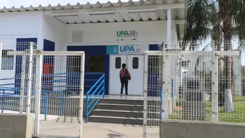 Prédio com entrada aberta e pessoa caminhando, cercado por cerca metálica e árvore.