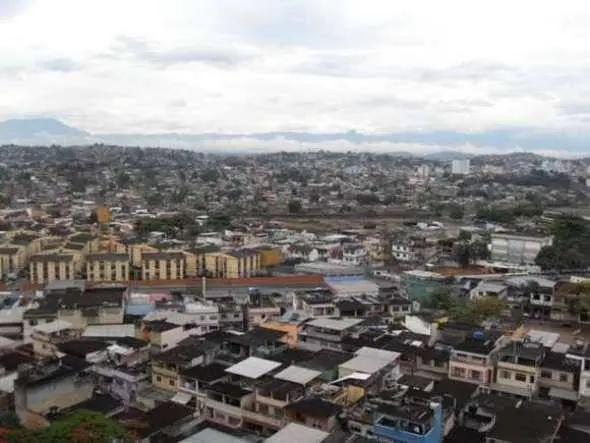Cidade com edifícios, ponte, árvores e montanhas cobertas por nuvens.