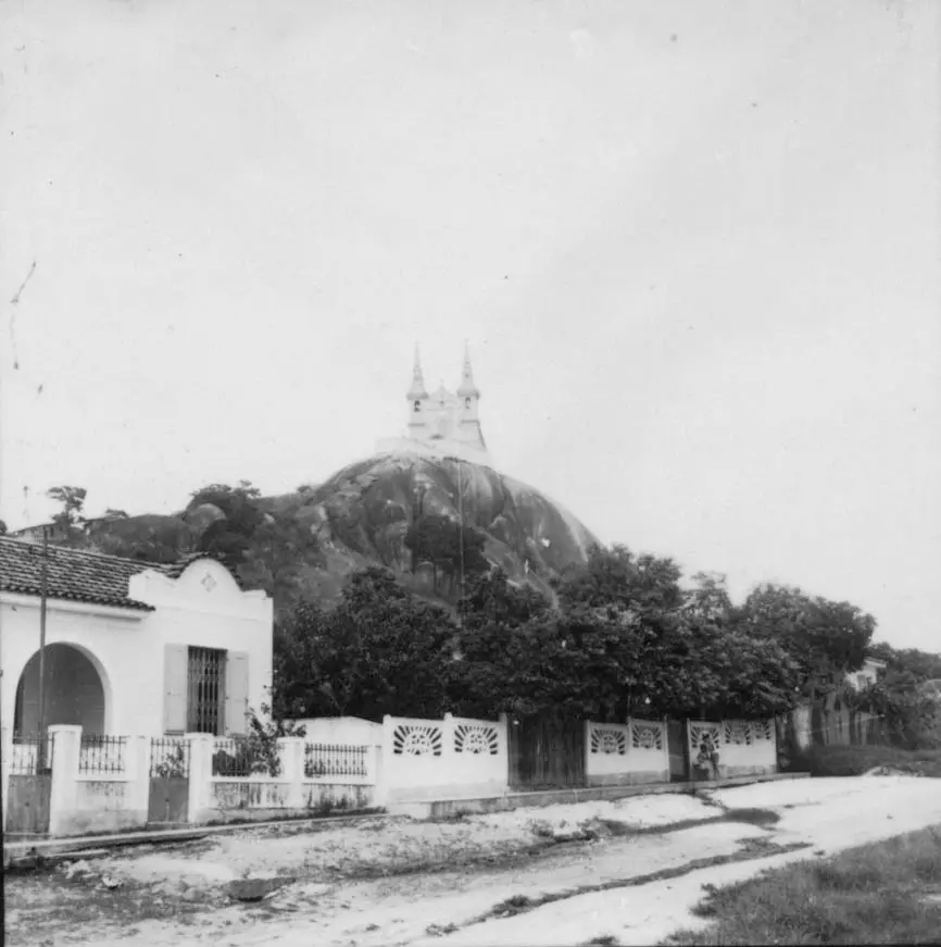 Cena urbana preto e branco com rocha, casa branca, cerca decorativa, área verde e estrutura com torres.