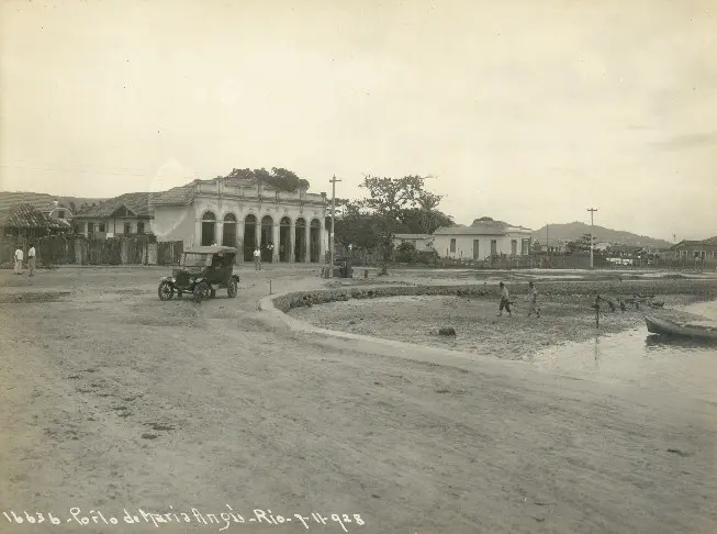 Uma praça antiga com edifícios, carro antigo e barco à beira-mar.