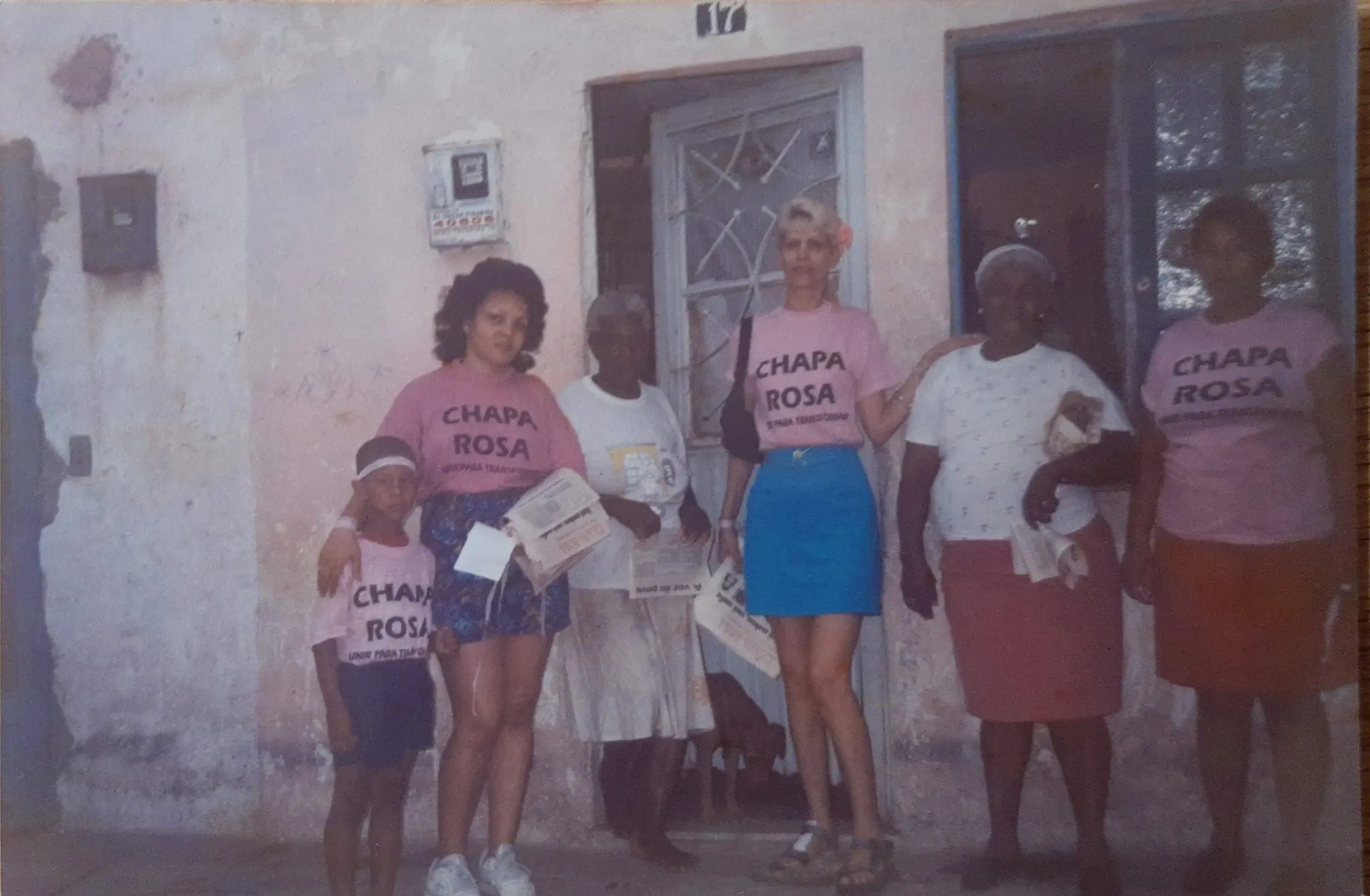 Pessoas posando em frente a casa com número 17, usando camisetas "Chapa Rosa".