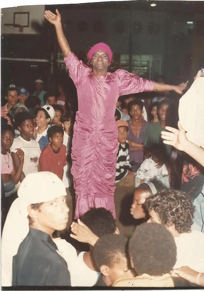 Pessoas em festa com vestido rosa e chapéu, multidão observando.