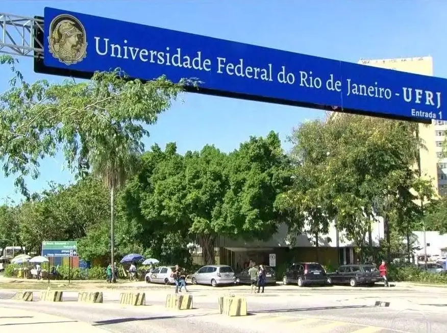 Campus universitário com árvores, sinal da UFRJ, prédios e céu azul.