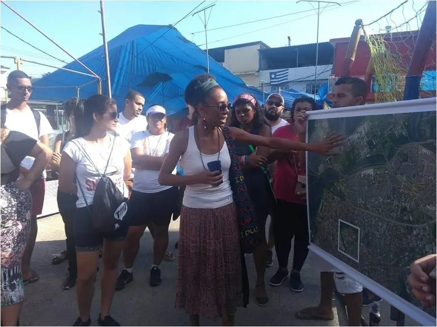 Grupo de pessoas reunidos ao ar livre, mulher centralizada aponta para um mapa.