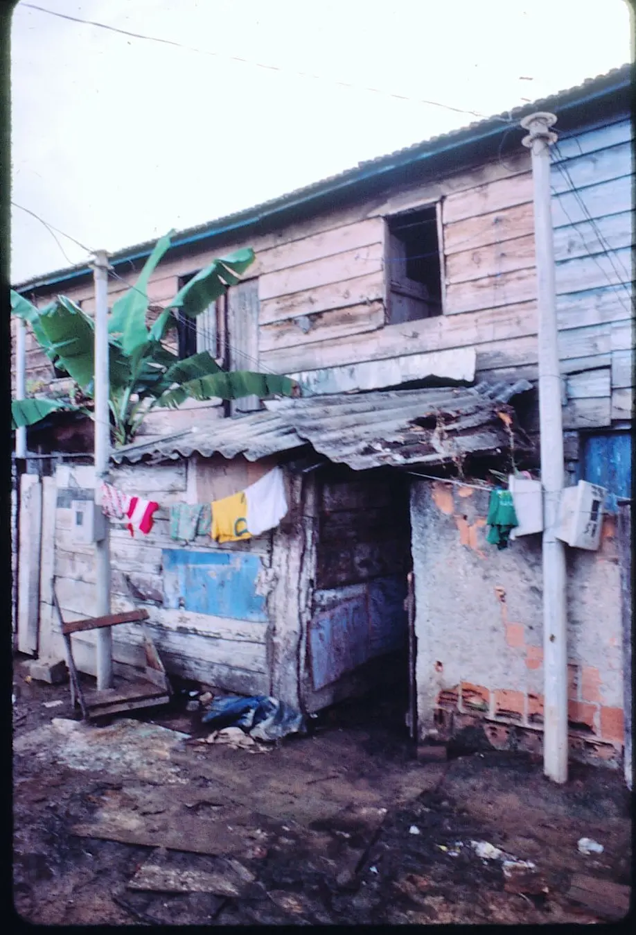Casa modesta em ambiente urbano, madeira, telhado inclinado, janelas abertas, planta verde, corda com roupas coloridas, caixa branca, chão de terra com madeira.