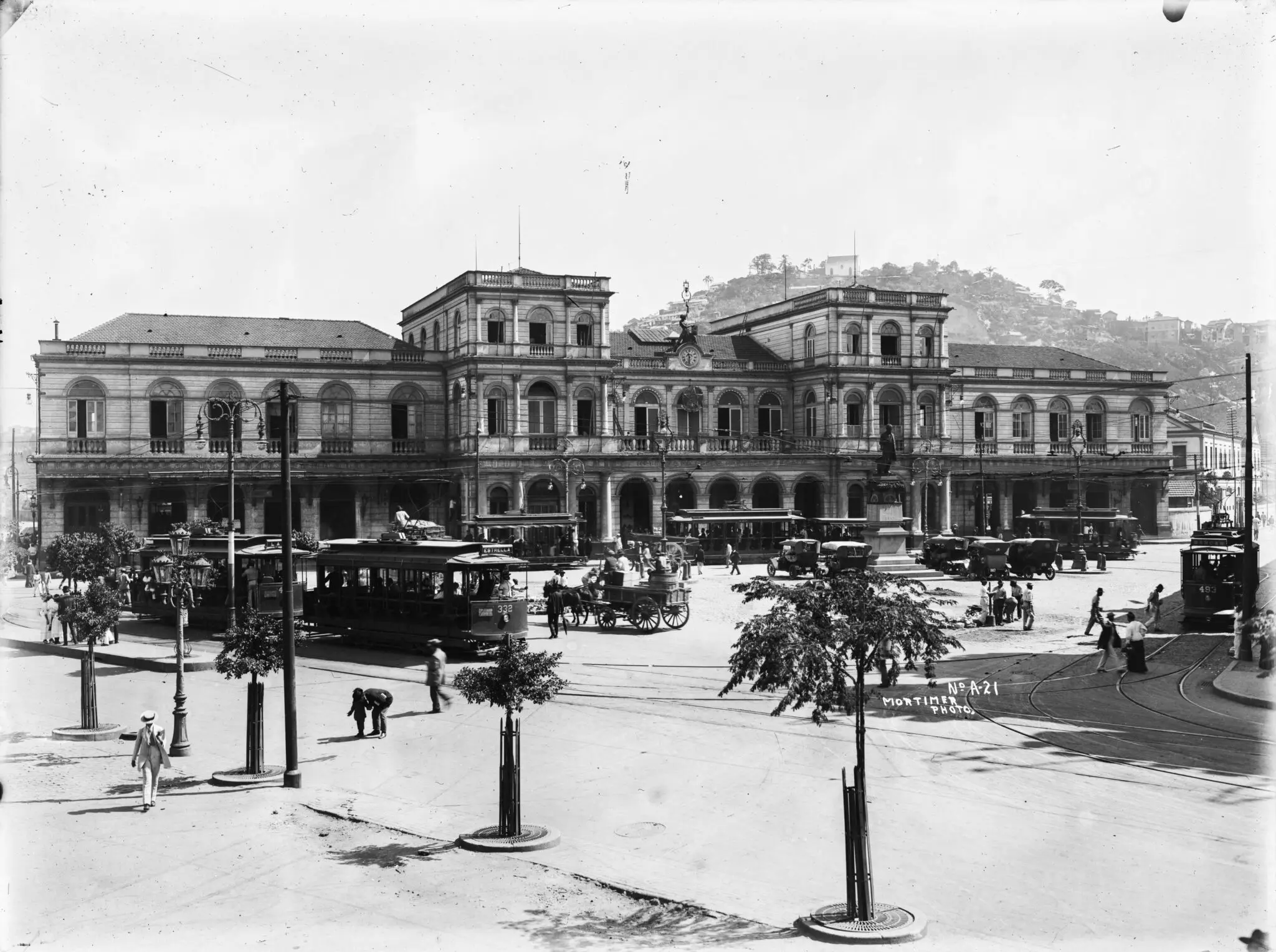 Edifício clássico em praça com árvores e pessoas caminhando.
