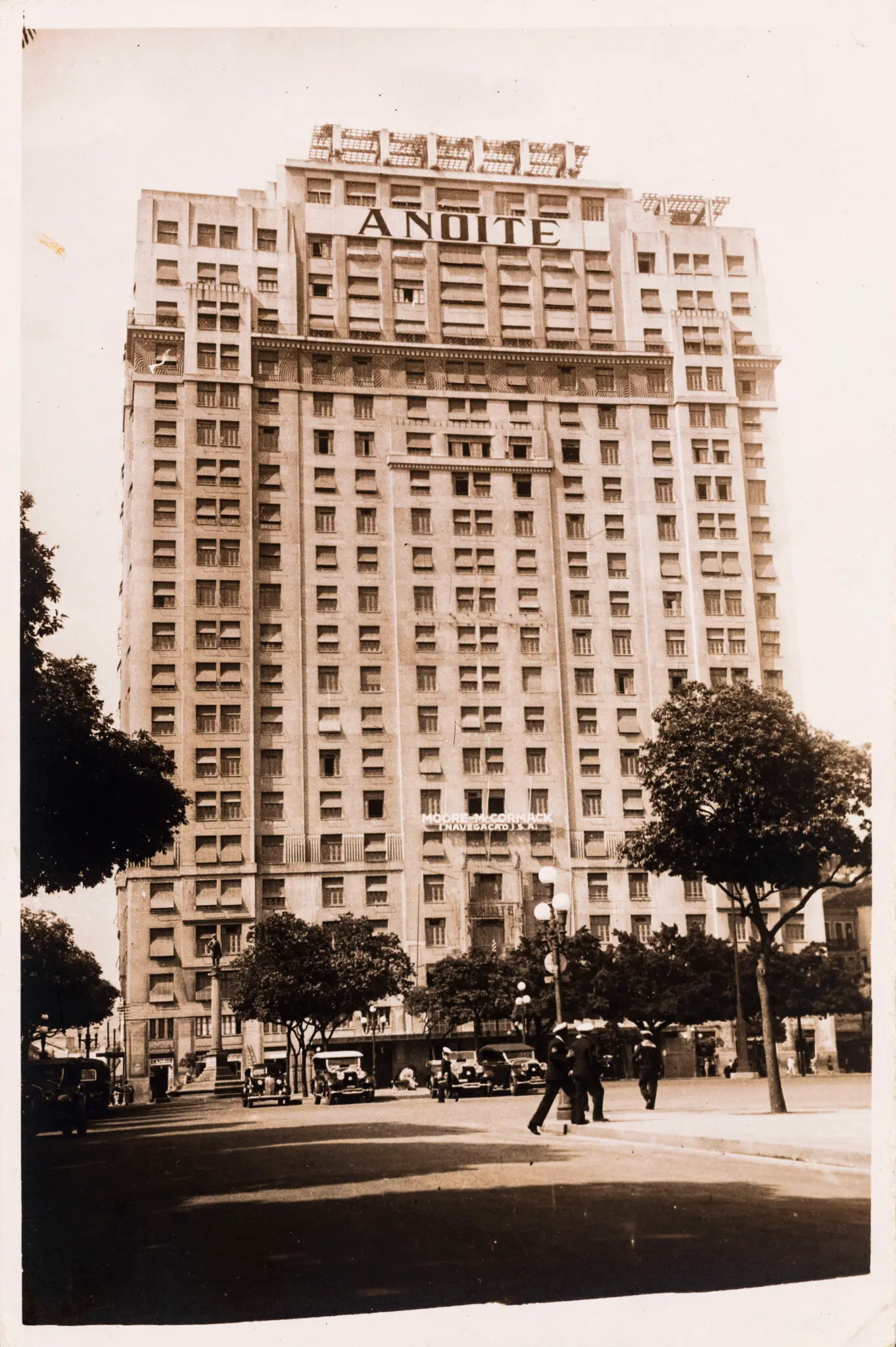 Edifício alto com "A NOITE" iluminado, árvores e pessoas caminhando.