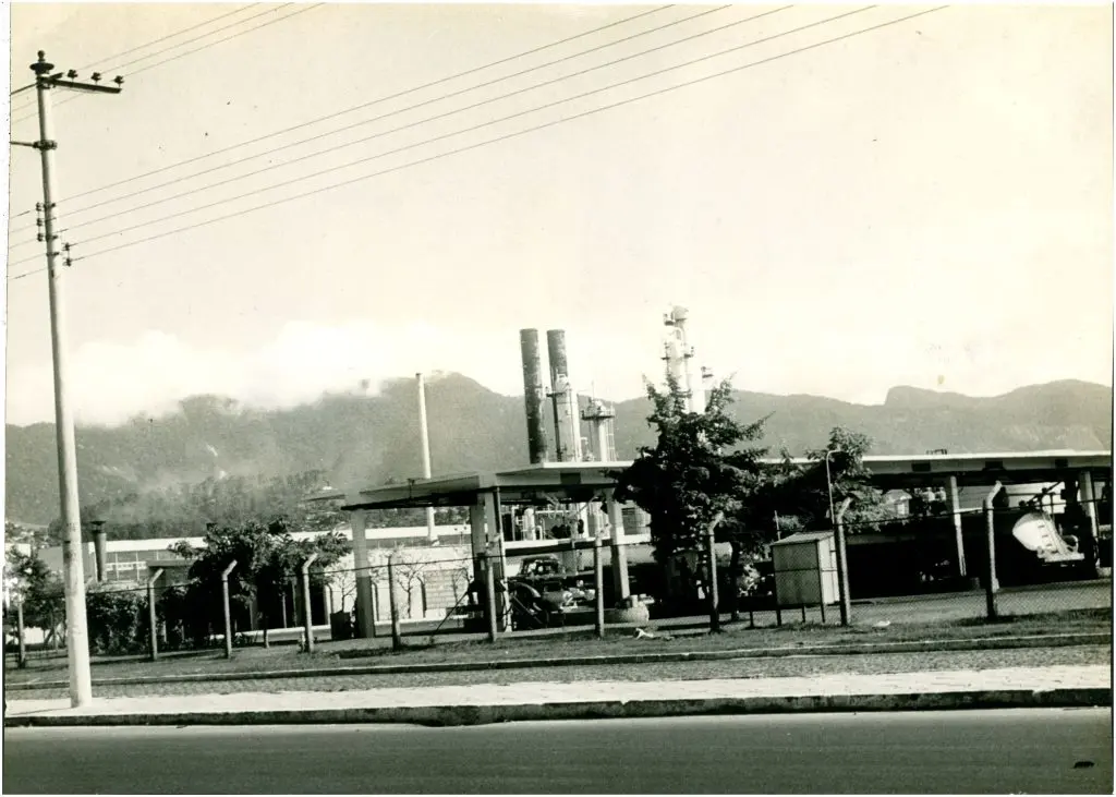 Área industrial com estruturas industriais, árvore e montanhas ao fundo.