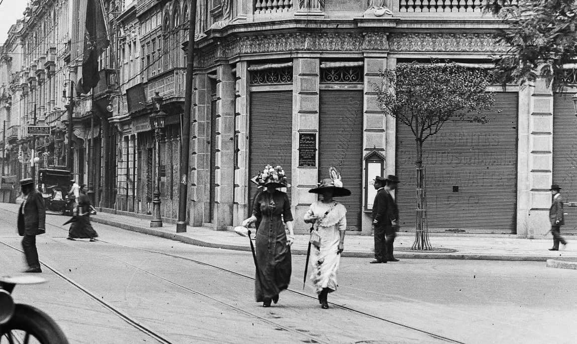 Rua urbana com edifícios históricos, carro antigo e pessoas caminhando.