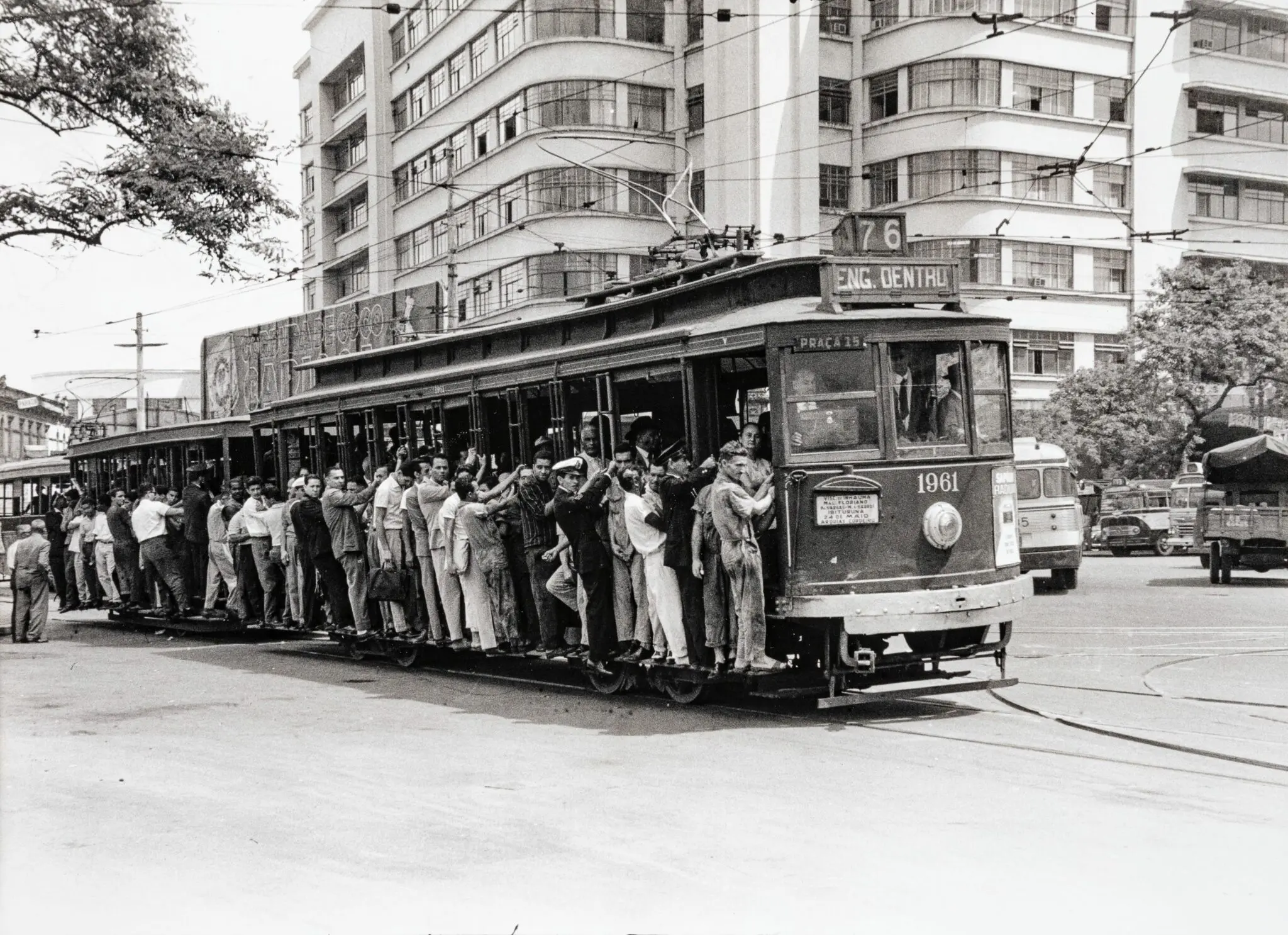 Bonde lotado em rua urbana, árvores e edifício ao fundo.