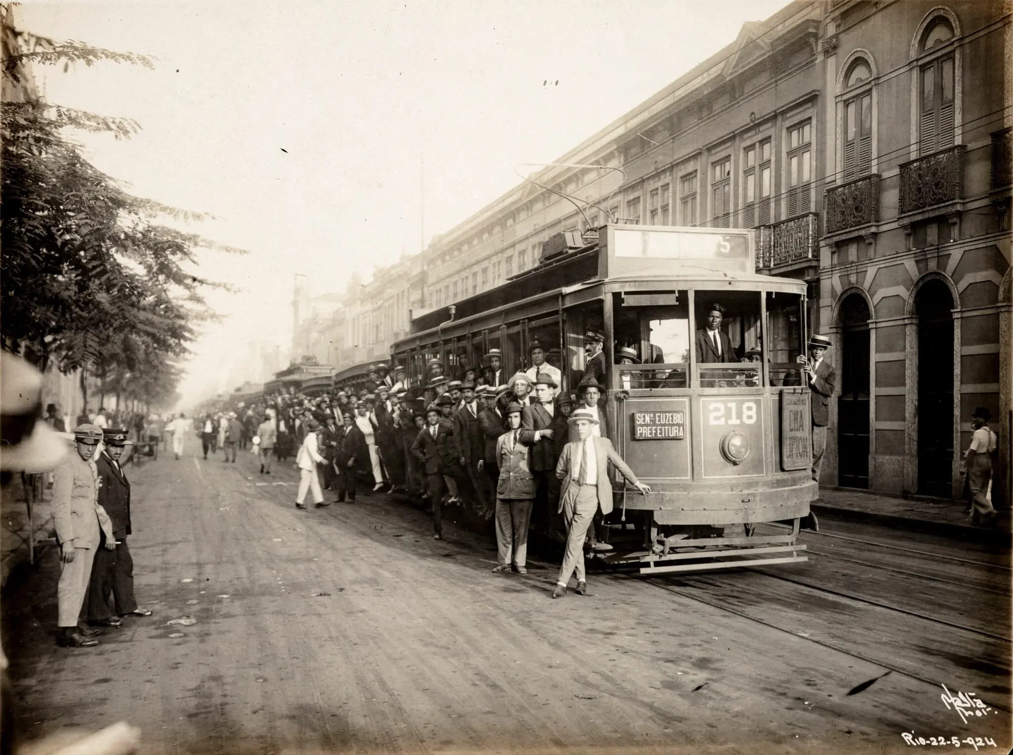 Bondinho antigo parado na rua, com árvores e edifício ao fundo.