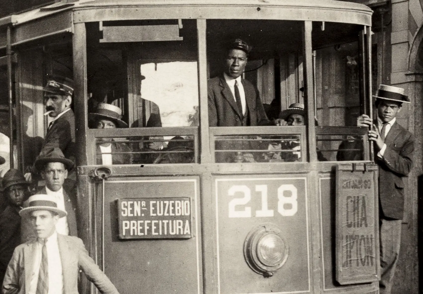 Grupo de pessoas em bonde preto e branco, motorista e passageiros.