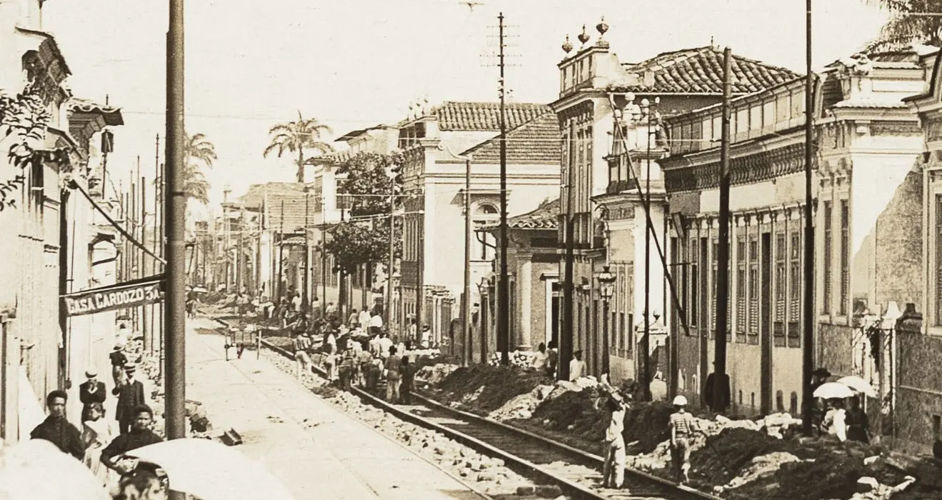 Uma rua urbana antiga em preto e branco, com construções antigas e pessoas caminhando.