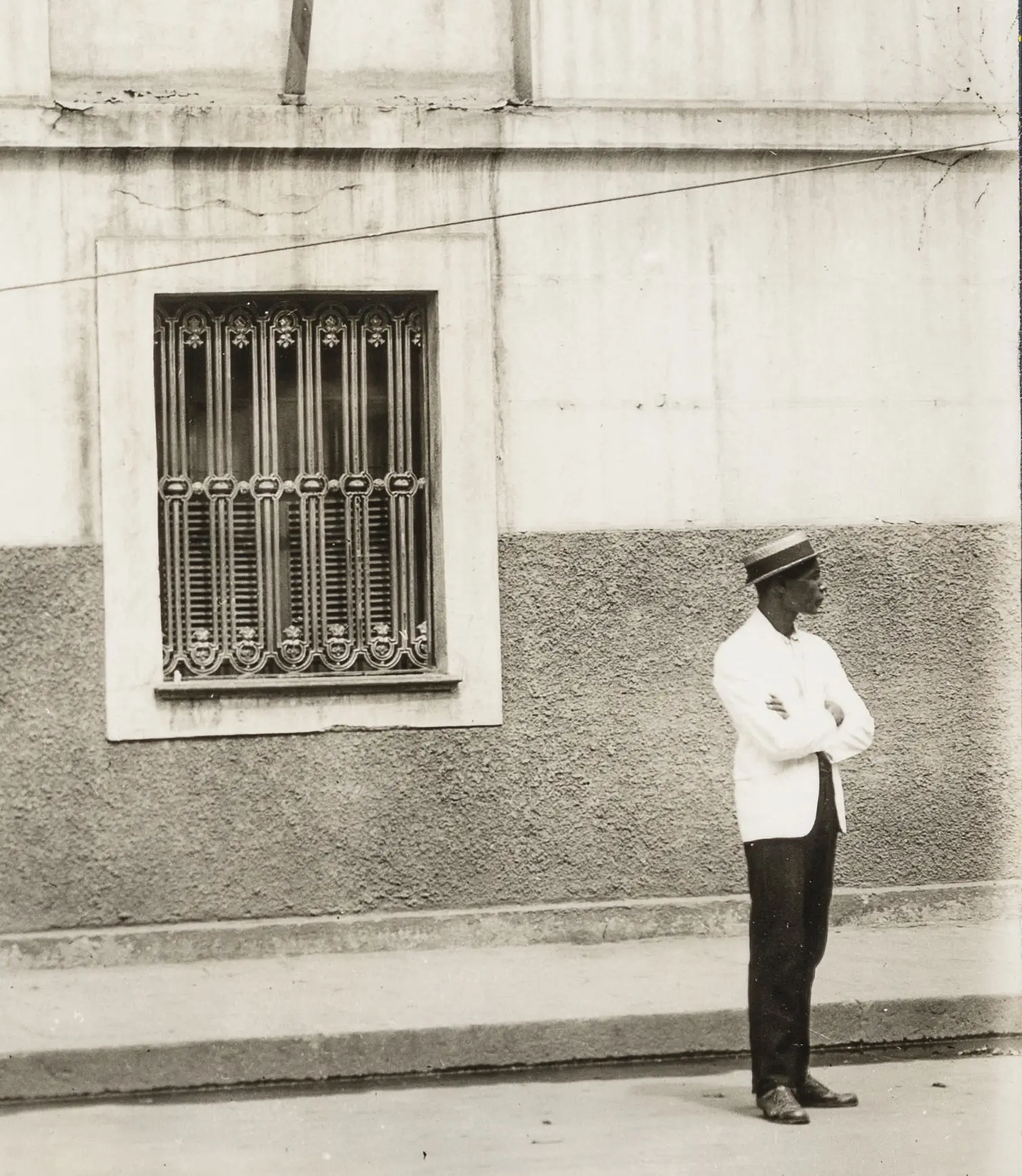 Homem de costas, chapéu e camisa branca, olhando para janela com grades metálicas em prédio preto e branco.