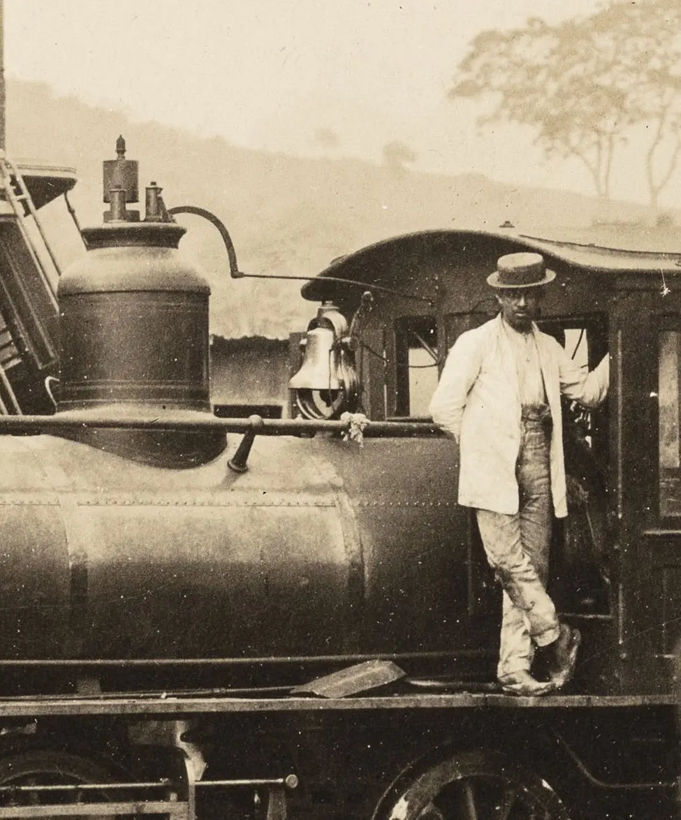 Homem em chapéu de palha perto de locomotiva antiga.