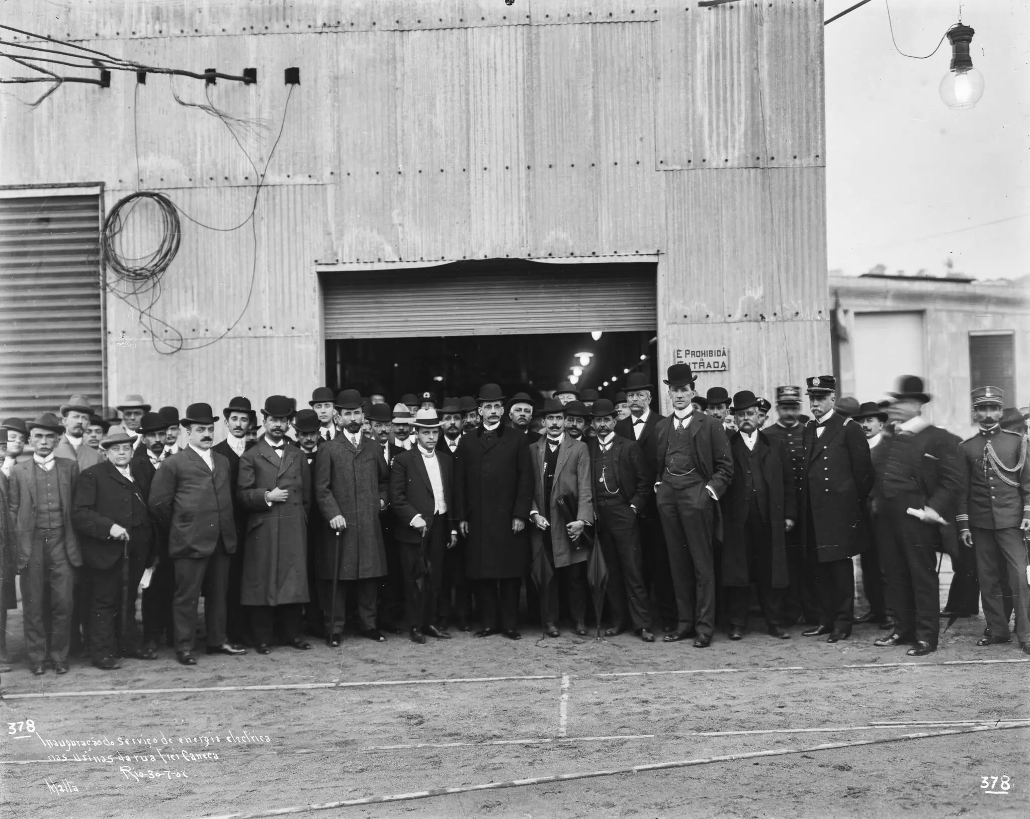 Grupo de pessoas em frente a edifício industrial, trajes formais do início do século XX.