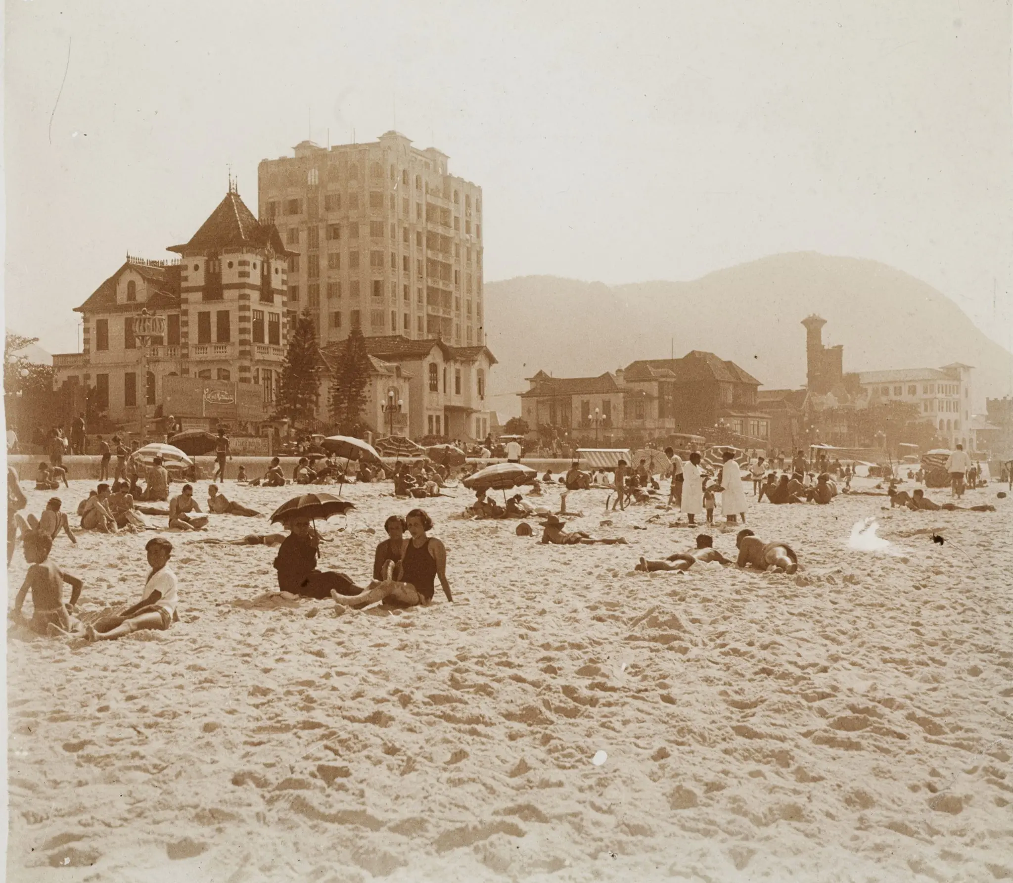 Praia lotada com edifícios e montanha ao fundo.
