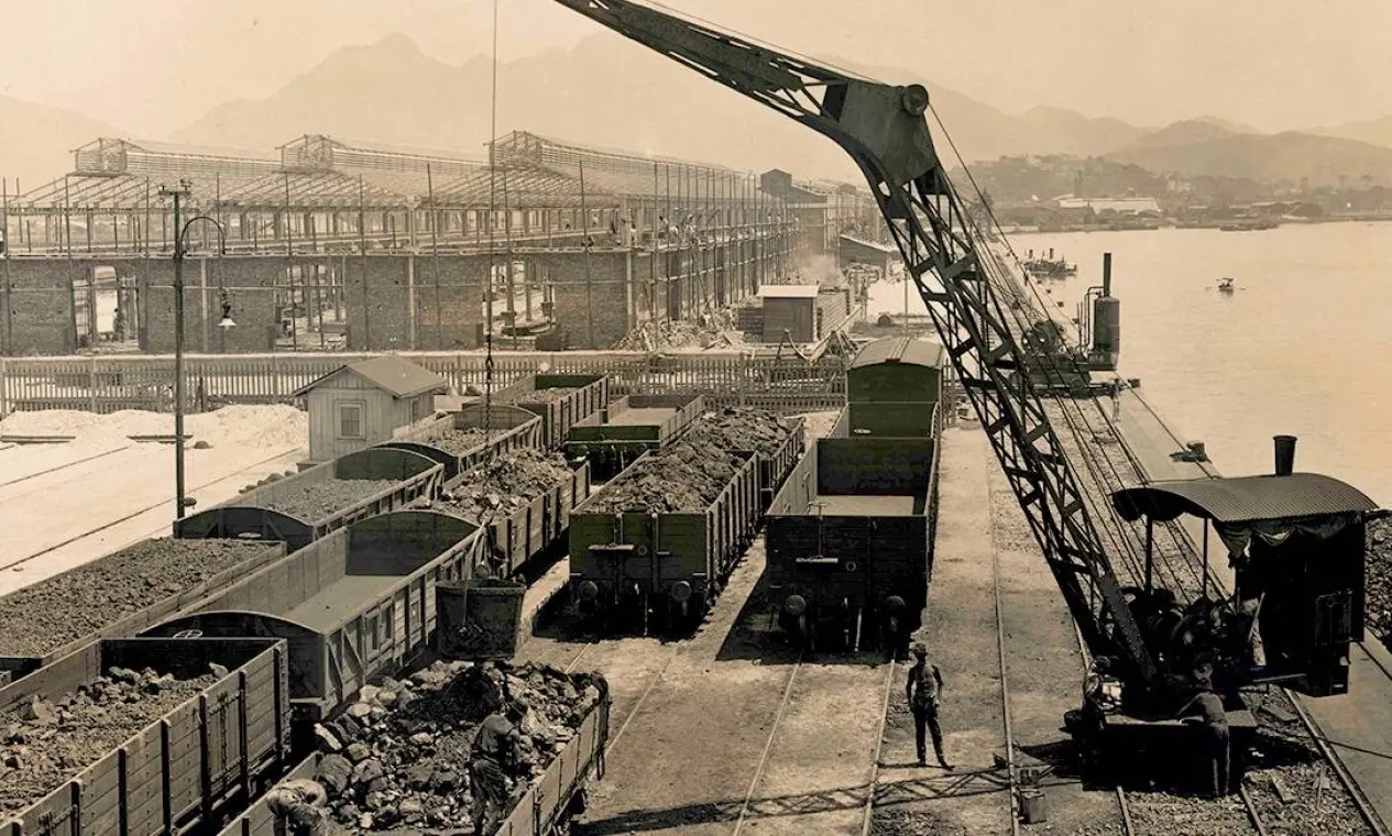 Porto antigo com vagões de trem, construção industrial, guindaste e casinha isolada.