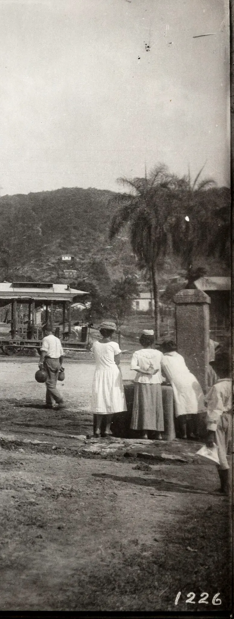 Fotografia preto e branco de uma cena urbana com bonde, homens caminhando, mulheres conversando e colina verde.