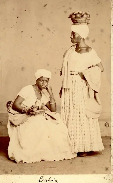 Duas mulheres em trajes tradicionais, uma sentada e outra de pé com chapéu.
