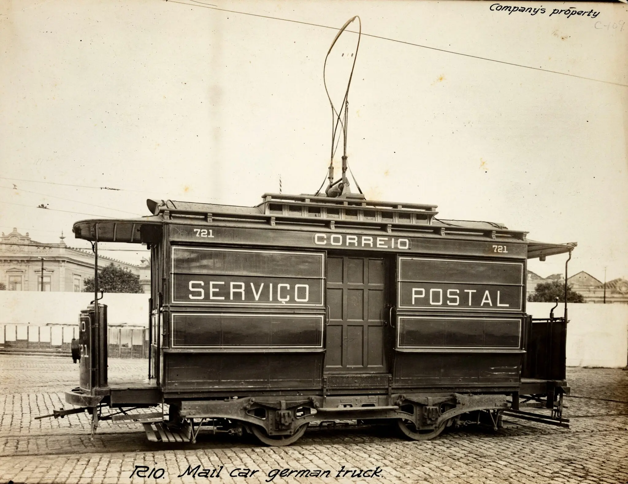 Bondinho elétrico antigo com inscrições "CORREIO" e "SERVIÇO POSTAL", num prédio clássico ao fundo.