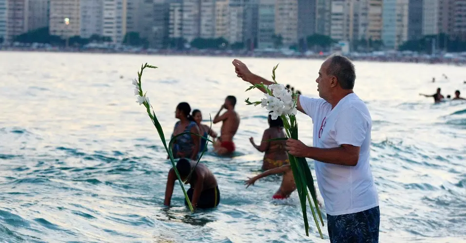 Praia com pessoas se divertindo no mar, homem segurando flores e folhas.
