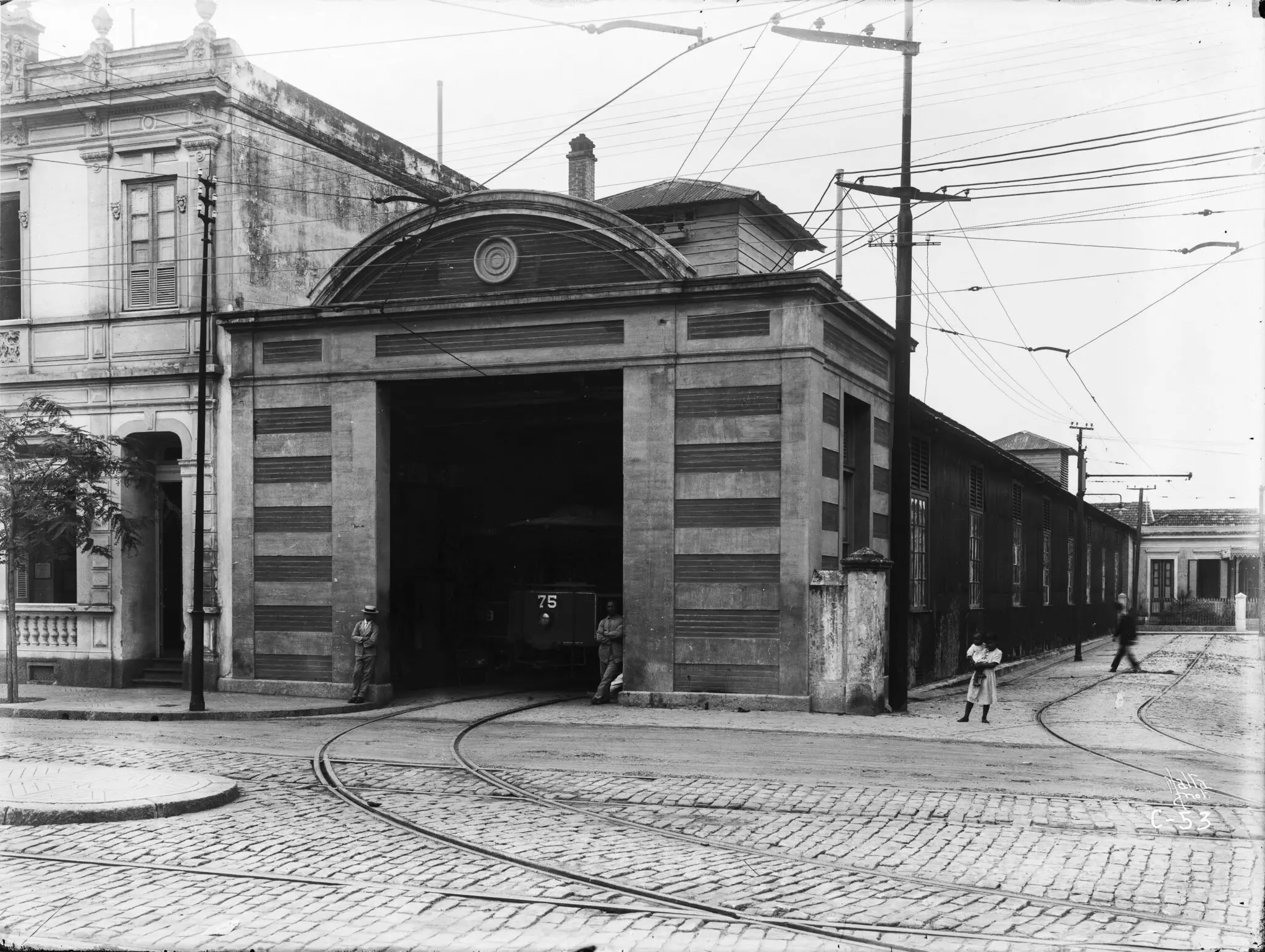 Garagem de veículos de transporte público com estrutura aberta, veículo visível, edifícios à volta e trilhas de trânsito.