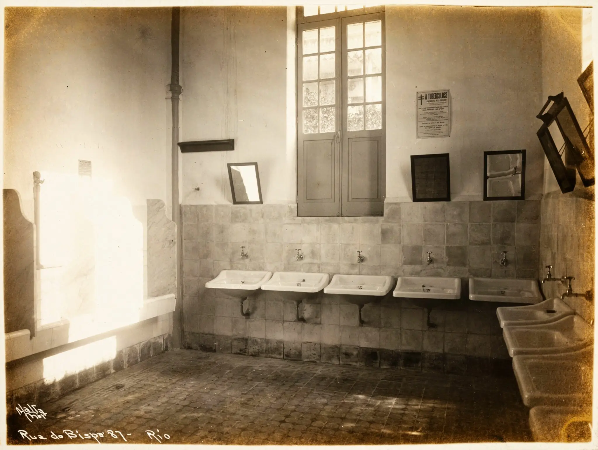 Banheiro público antigo com cinco lavabos, janela, porta dupla, arte, espelho e azulejos.