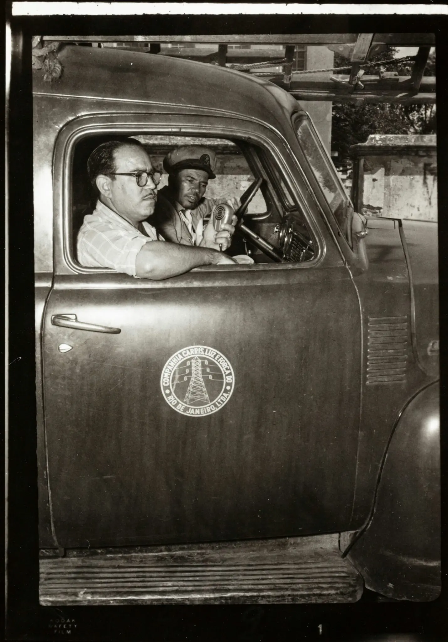 Dois homens em carro antigo, preto e branco.