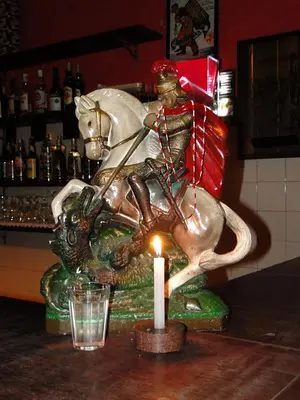 Estátua de São Jorge em bar/restaurante, vela acesa, barra com bebidas.