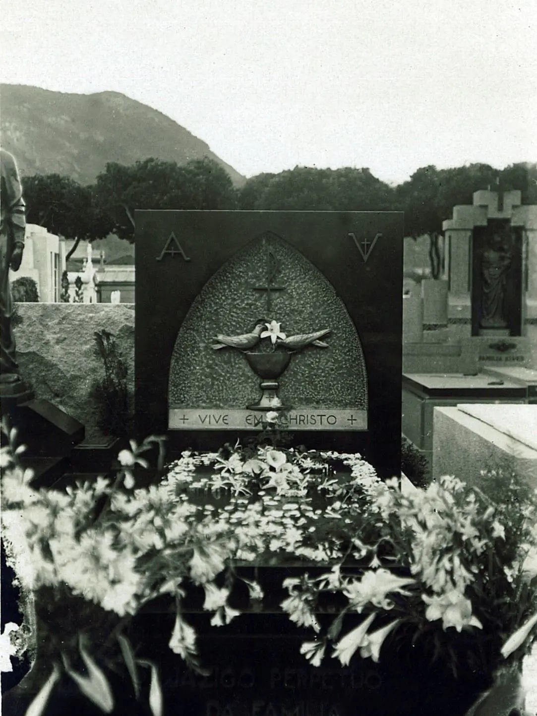 Monumento funerário com inscrição "VIVE EM CRISTO", flores brancas e pombos.