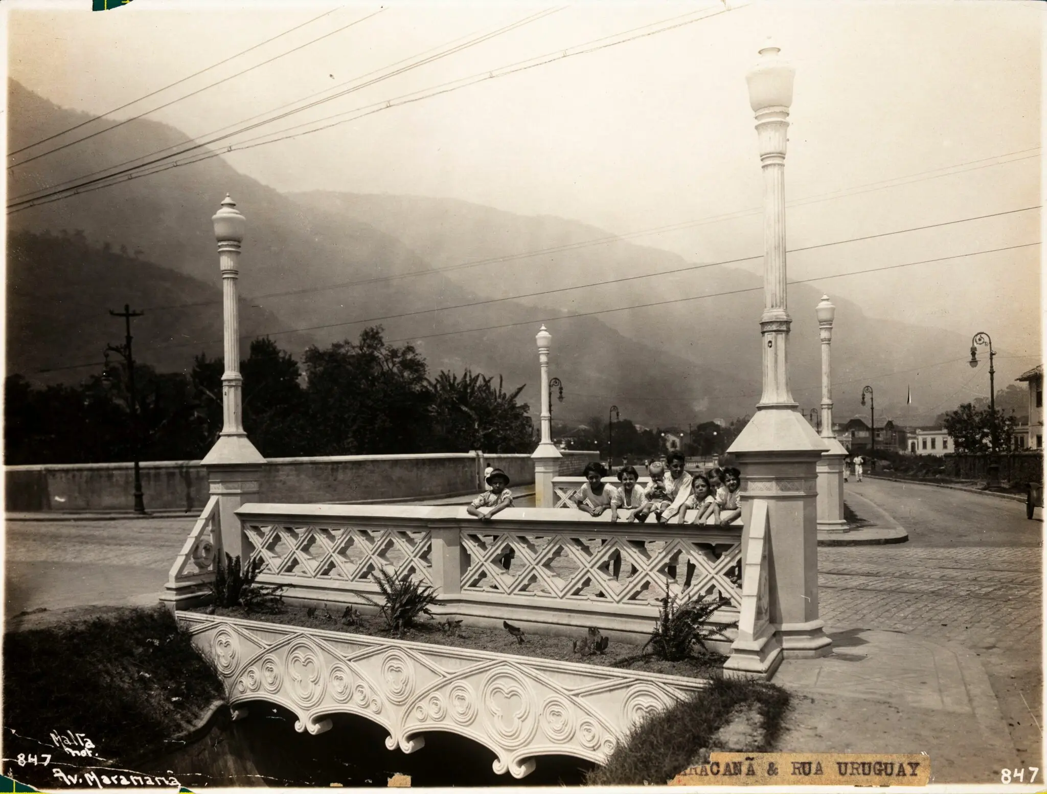 Cenário urbano com ponte de pedra, crianças sentadas, montanhas ao fundo.