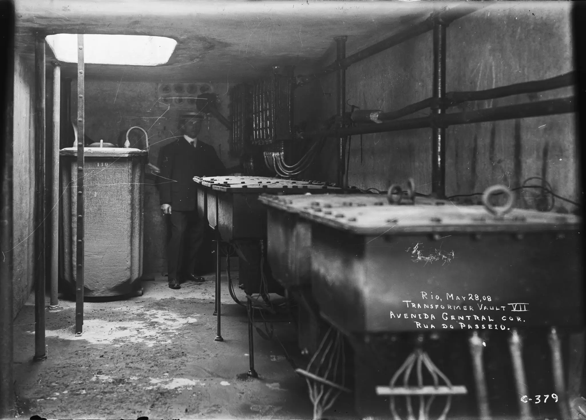 Interior de câmara de transformadores com homem observando, tanques metálicos à direita.