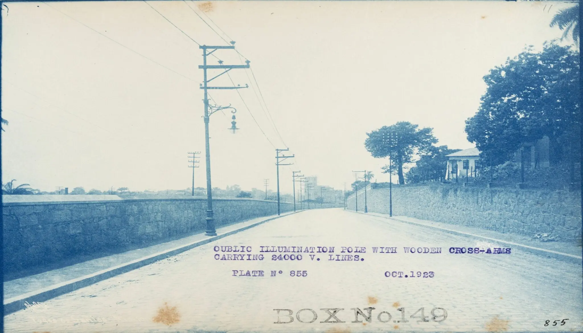Fotografia antiga de rua com muro de pedra e postes de iluminação elétrica.