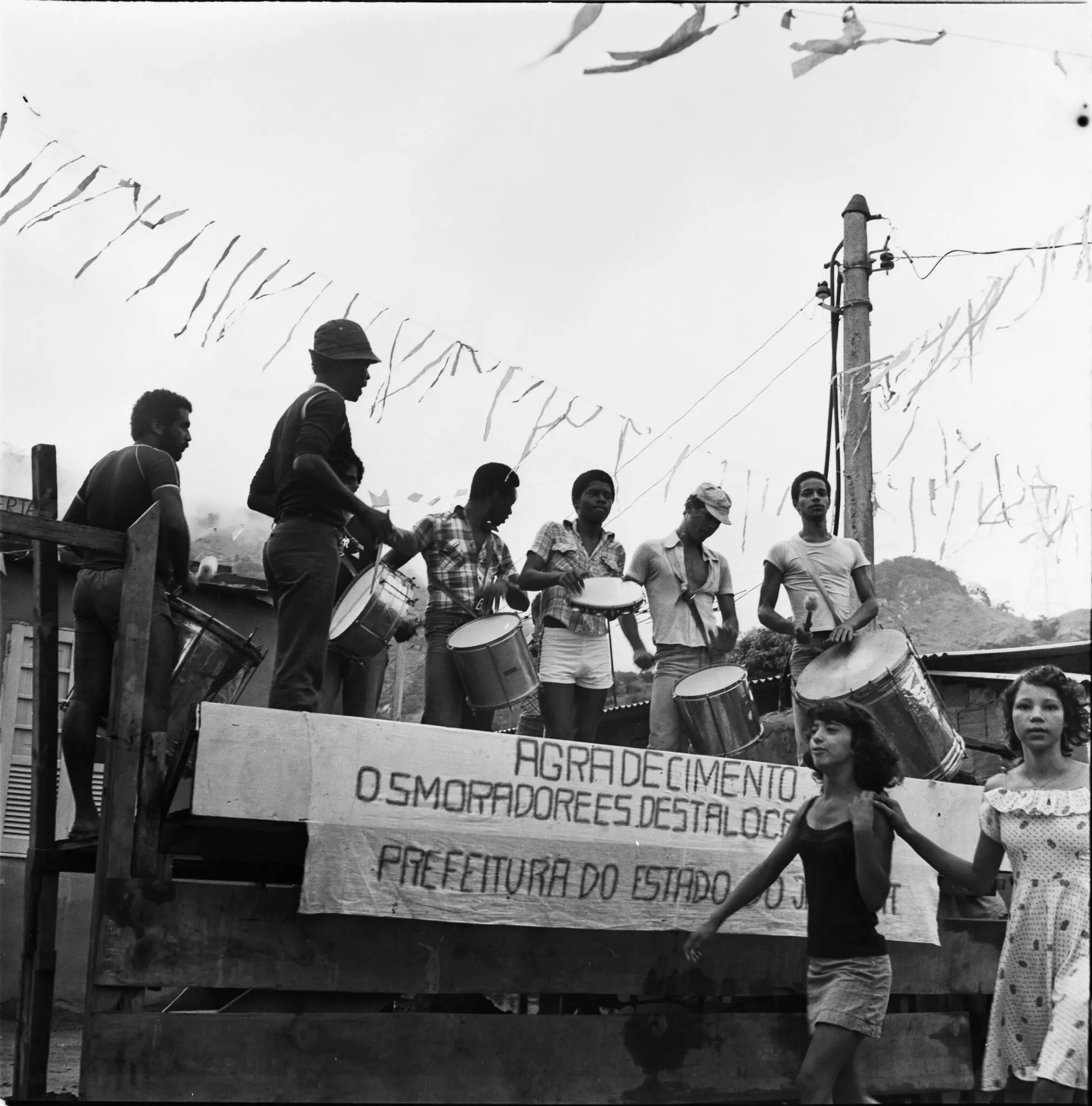 Pessoas tocando instrumentos em palco, antena e casas ao fundo.
