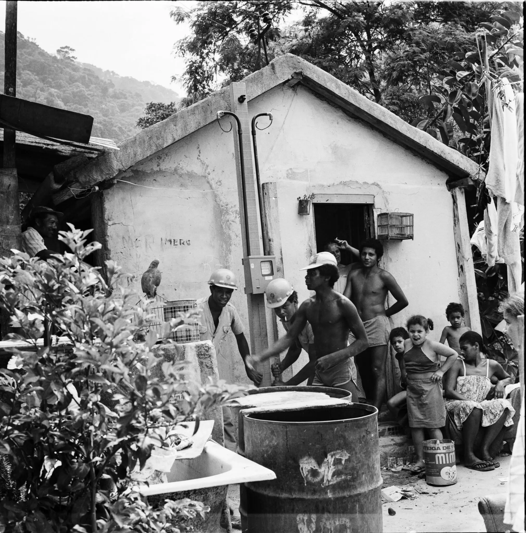 Cena de distribuição de energia em comunidade, com planta verde, casa branca, pessoas usando capacetes e crianças sentadas.