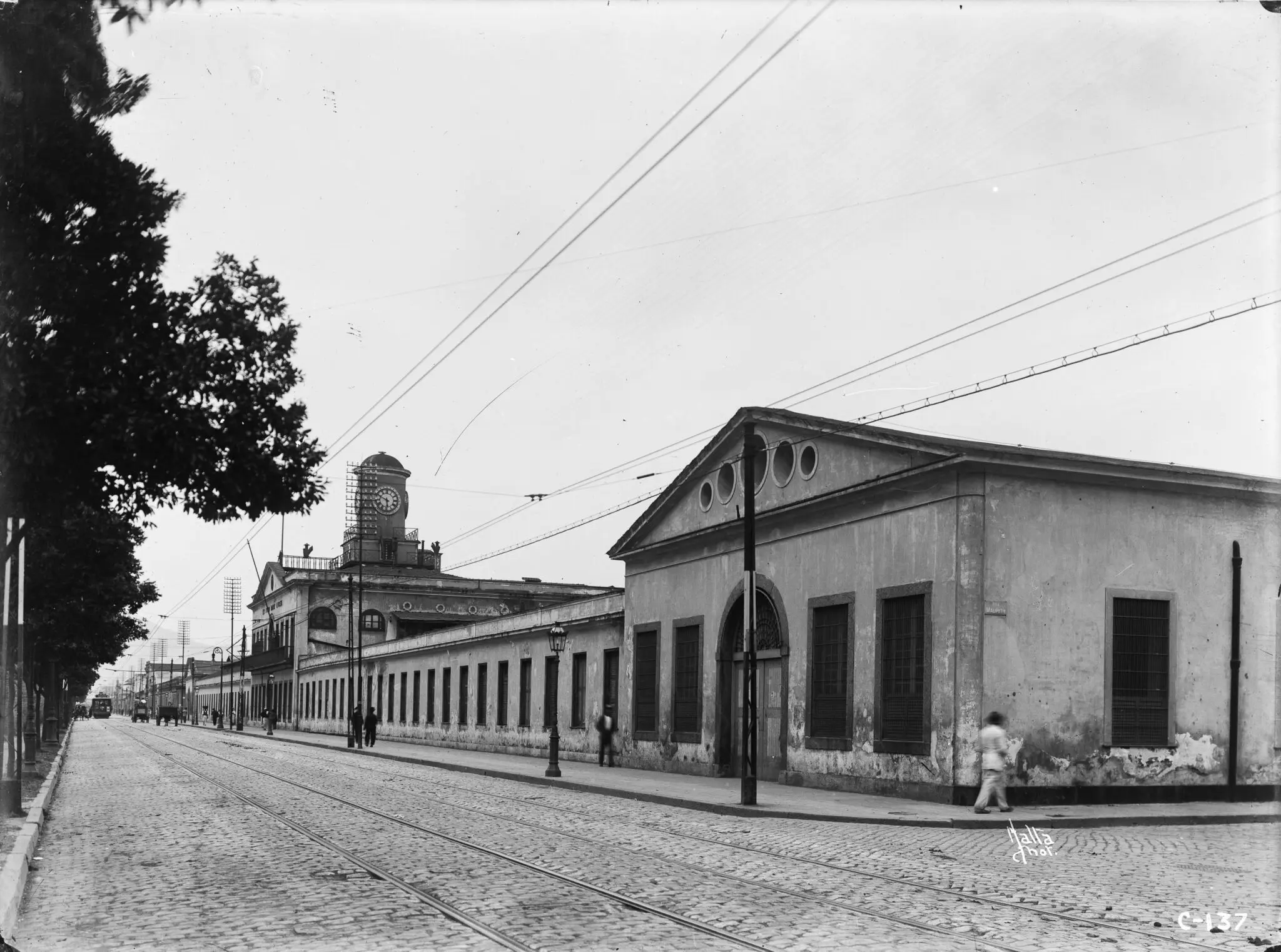 Fábrica de gás em Rio Antigo, com árvore verde, trilhos de bondes, construções e pessoas caminhando.