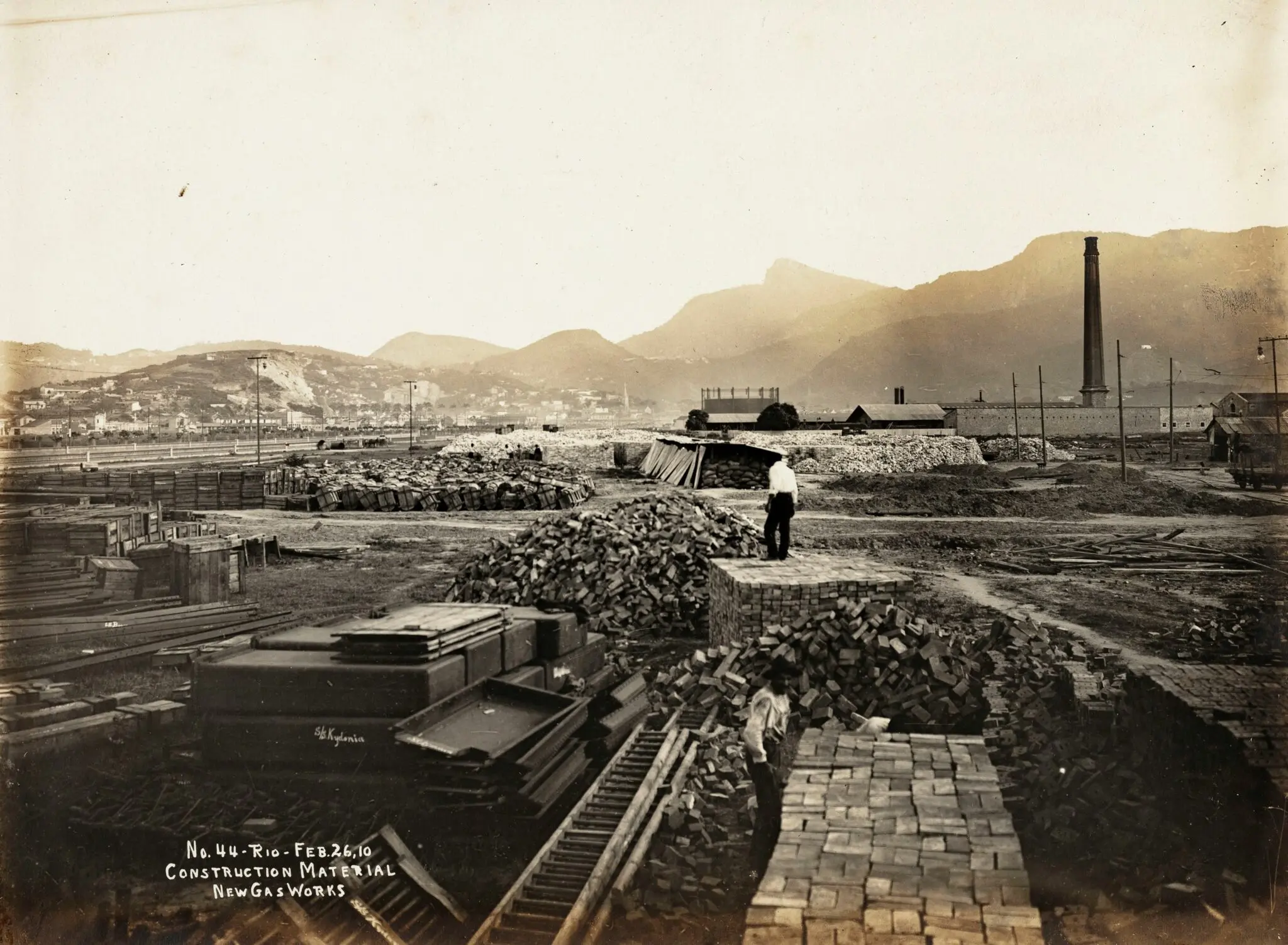 Fotografia antiga de construção com pilhas de madeira, tijolos e homem caminhando.