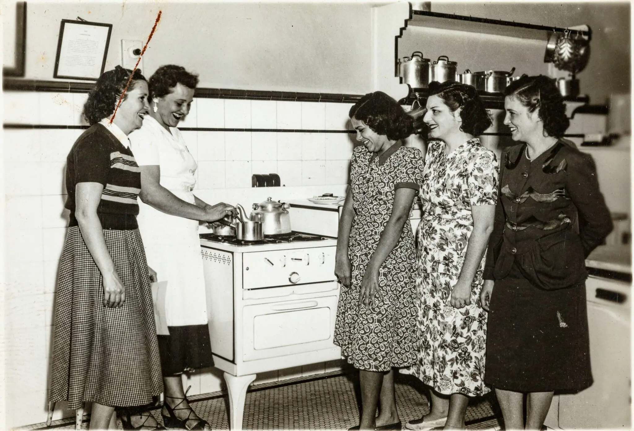 Mulheres em cozinha, conversando e sorrindo, com panela e utensílios ao fundo.