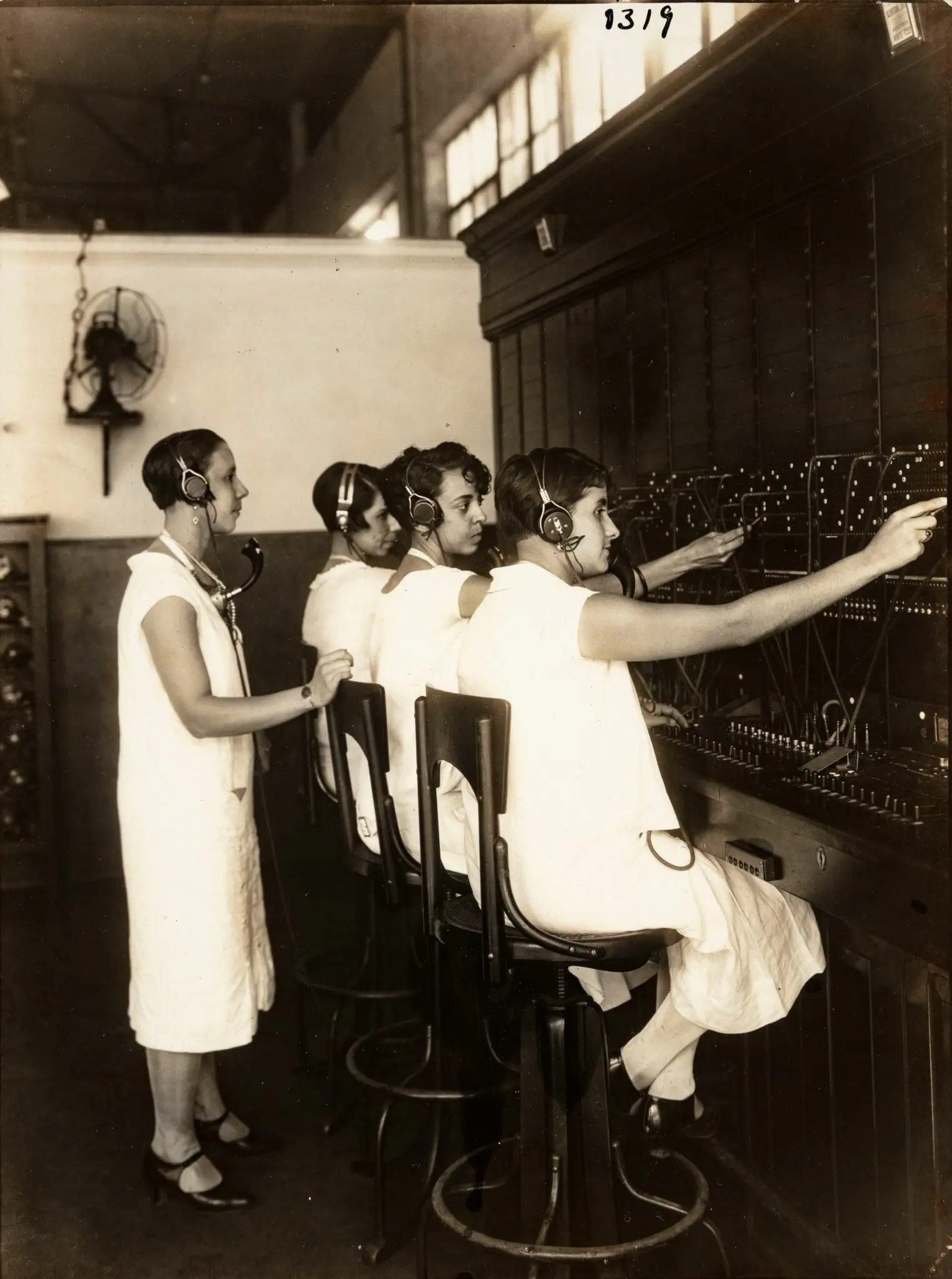 Quatro telefonistas em sala de operações, uniformes brancos, painel de controles.