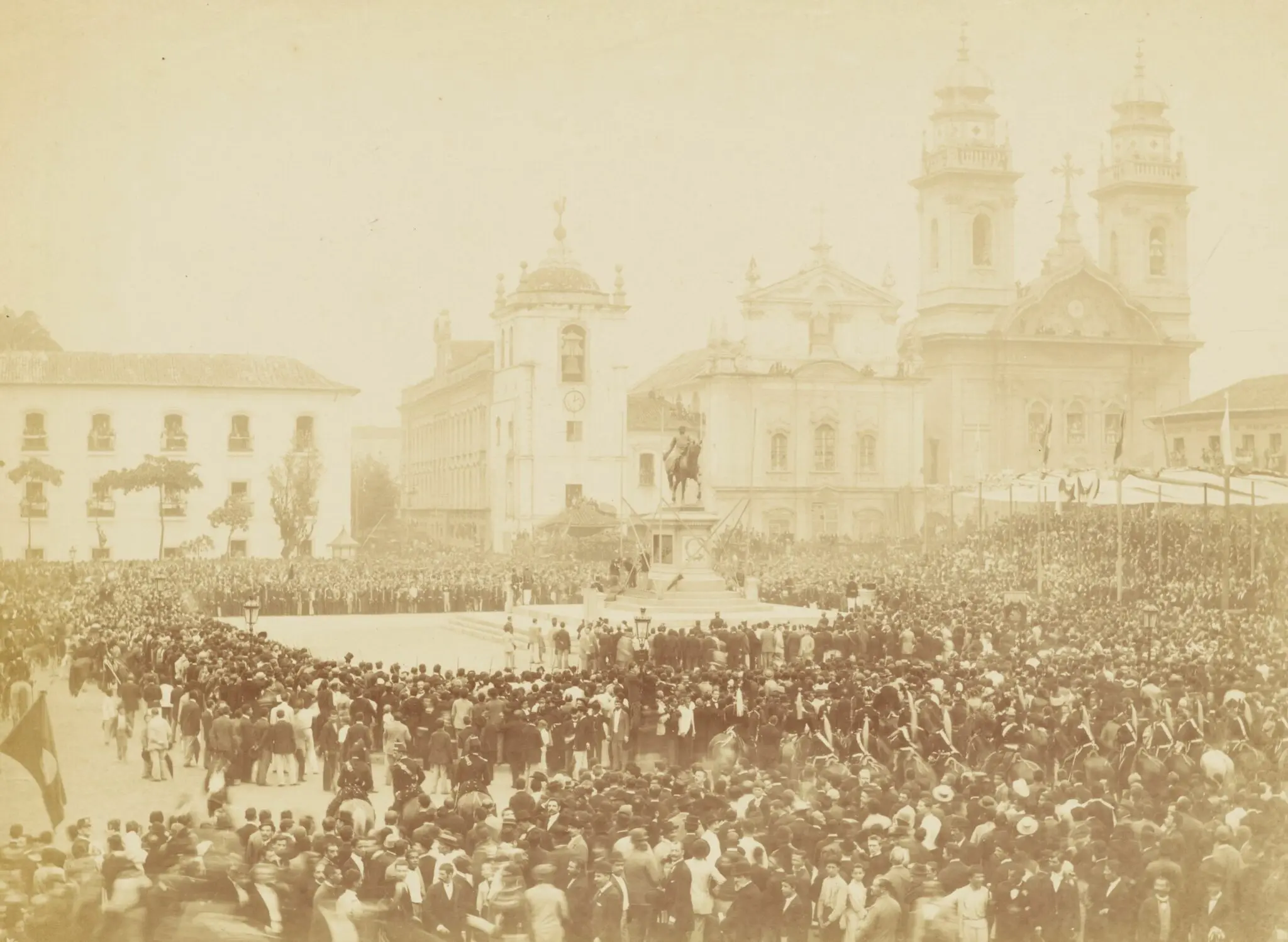 Multidão em praça histórica com estátua de general e igrejas ornamentadas.