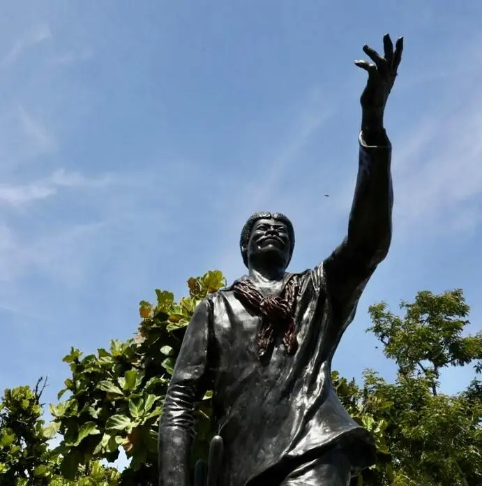Estátua de bronze com figura humana erguida, lenço colorido, árvores verdes e céu azul claro.