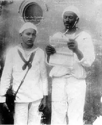 Dois marinheiros em uniforme segurando objetos e um papel em frente a uma janela com símbolo.