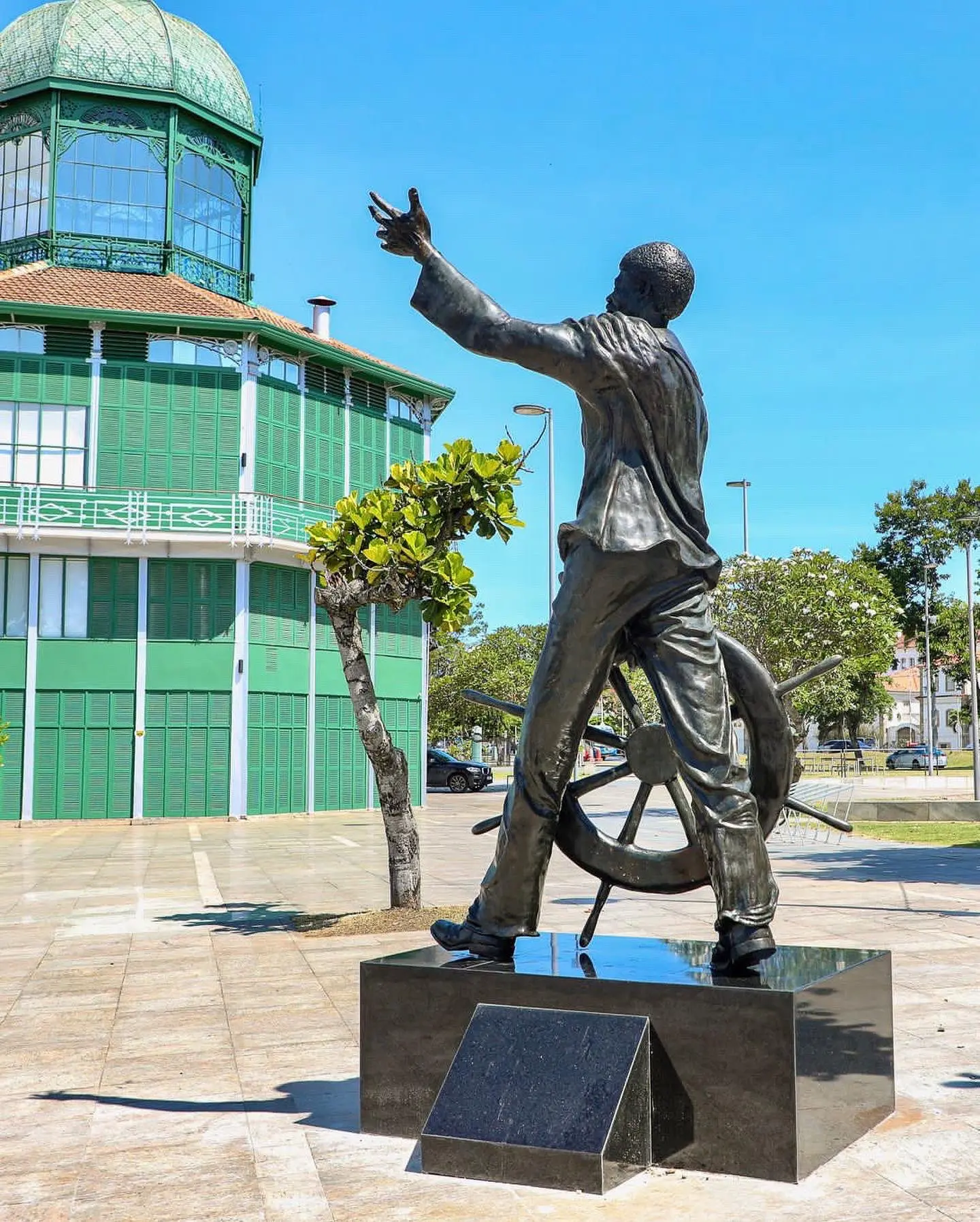 Estátua dinâmica em praça pública, com edifício verde ao fundo.